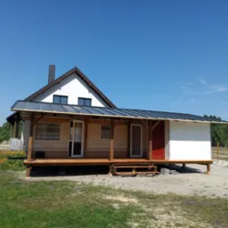 Dom z dobudowanym drewnianym tarasem zadaszonym blachą, widoczne schody i czerwone drzwi wejściowe, na trawniku kwiaty.