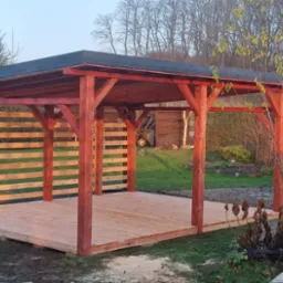 Nowoczesna, otwarta konstrukcja z drewna w kolorze czerwieni, z ażurowymi ścianami i płaskim dachem, na tle ogrodu z grillem.