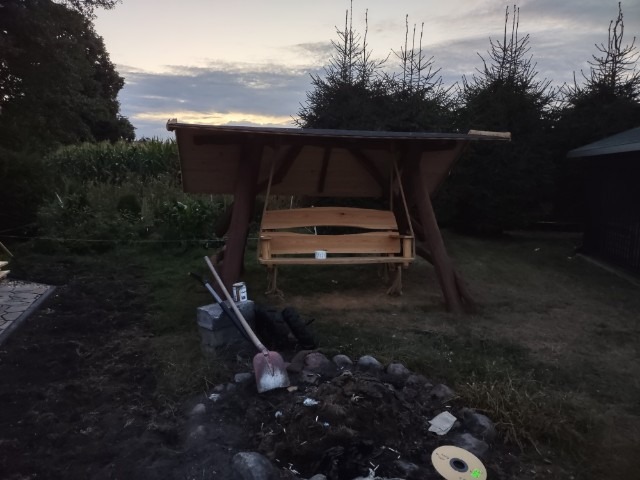 Drewniana huśtawka ogrodowa z zadaszeniem, widoczna podczas montażu, obok narzędzia i wykopana ziemia.