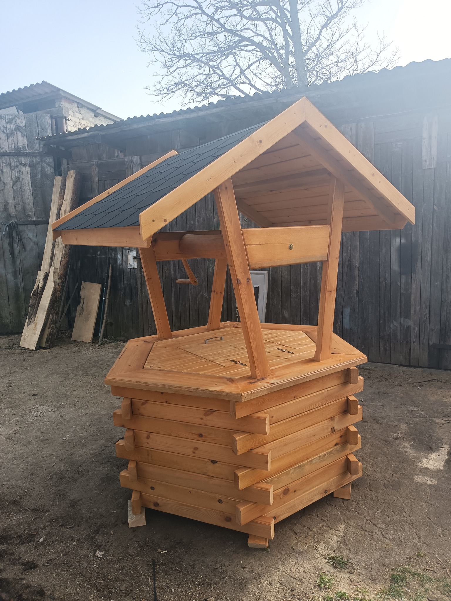 Drewniana studnia ogrodowa z czarnym dachem, wykonana z jasnego drewna, na tle szarego podwórza.