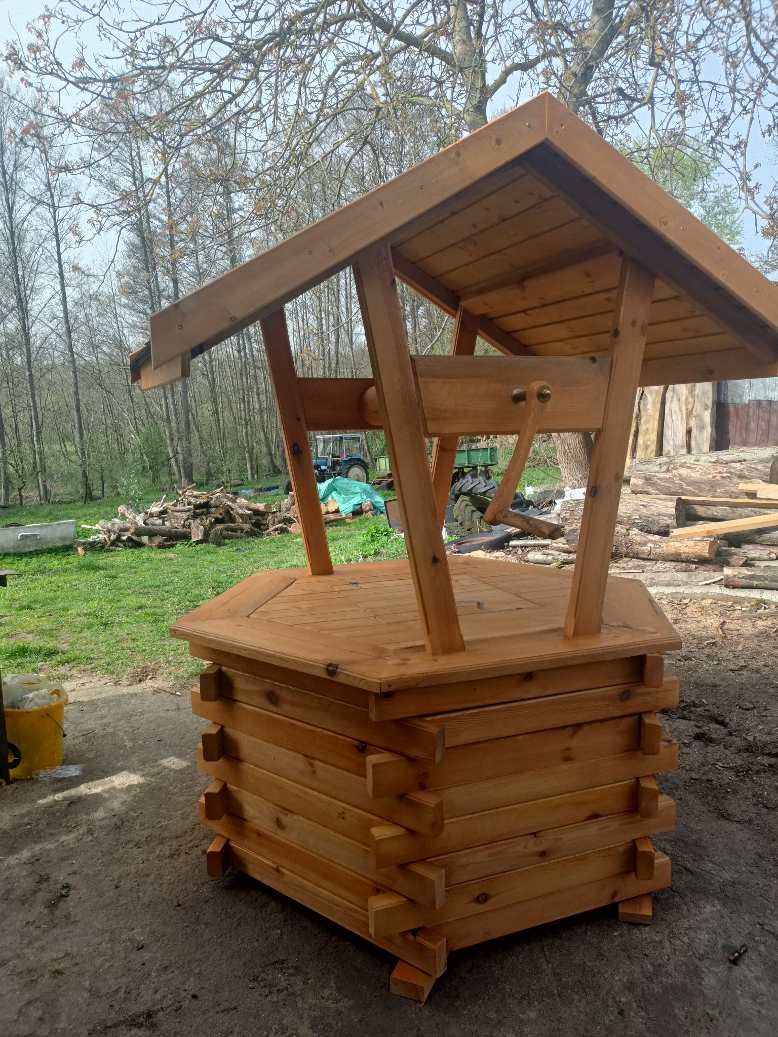 Studnia drewniana z daszkiem, wykonana z jasnego drewna, na tle trawy, drzew i sterty pociętego drewna.