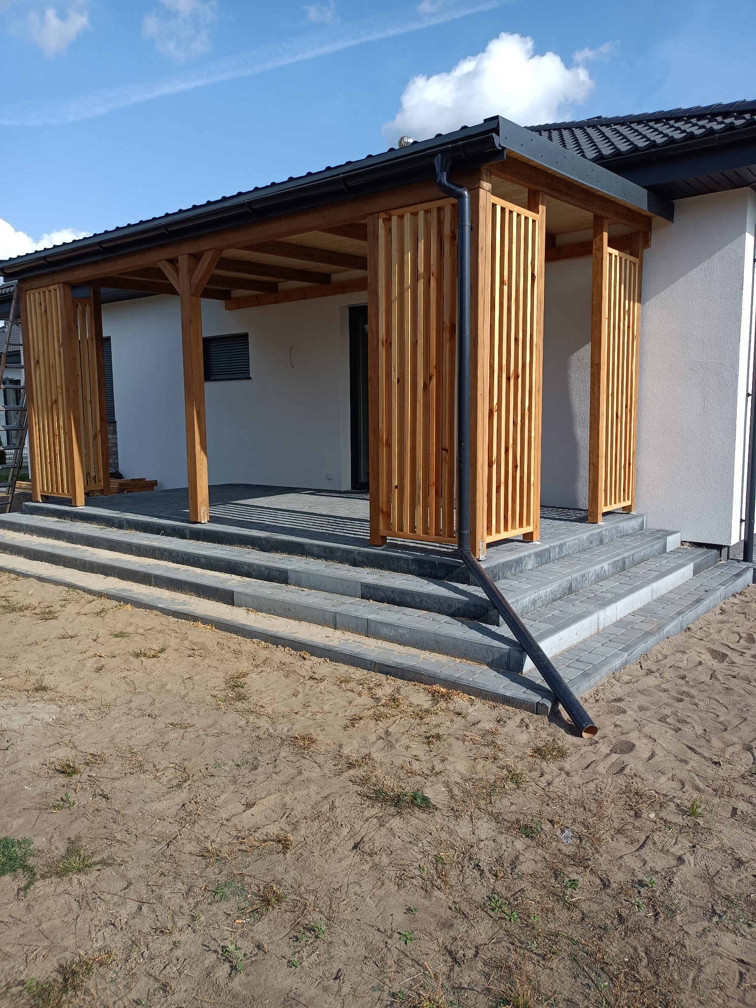 Drewniana pergola z pionowymi lamelami przy nowoczesnym domu, z widocznymi schodami z kostki brukowej i rynną spustową.