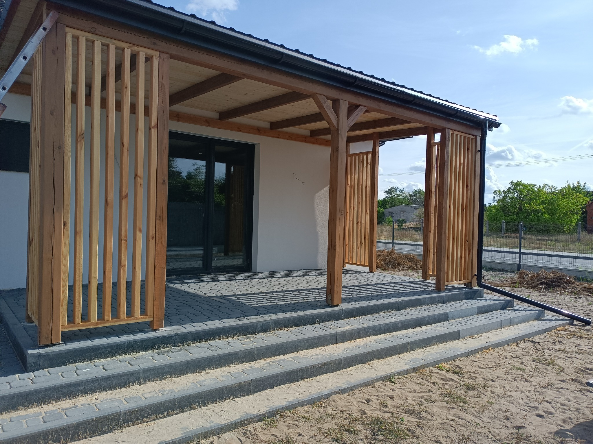 Nowoczesna pergola z drewnianymi lamelami, wsparta na kamiennych schodach, z widokiem na ogród w tle.