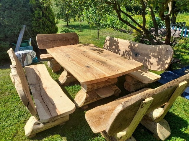 Rustykalny zestaw mebli ogrodowych z litego drewna: stół z grubym blatem i ławy z oparciami, wykonane z surowych bali, na tle zielonego trawnika i drzew.