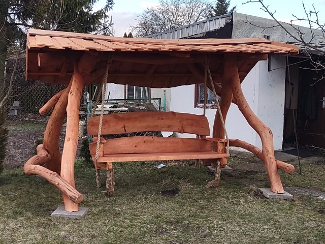 Drewniana huśtawka ogrodowa z dachem, wykonana z naturalnych pni drzew, zawieszona na grubych linach, z widocznym tłem budynku i zieleni.