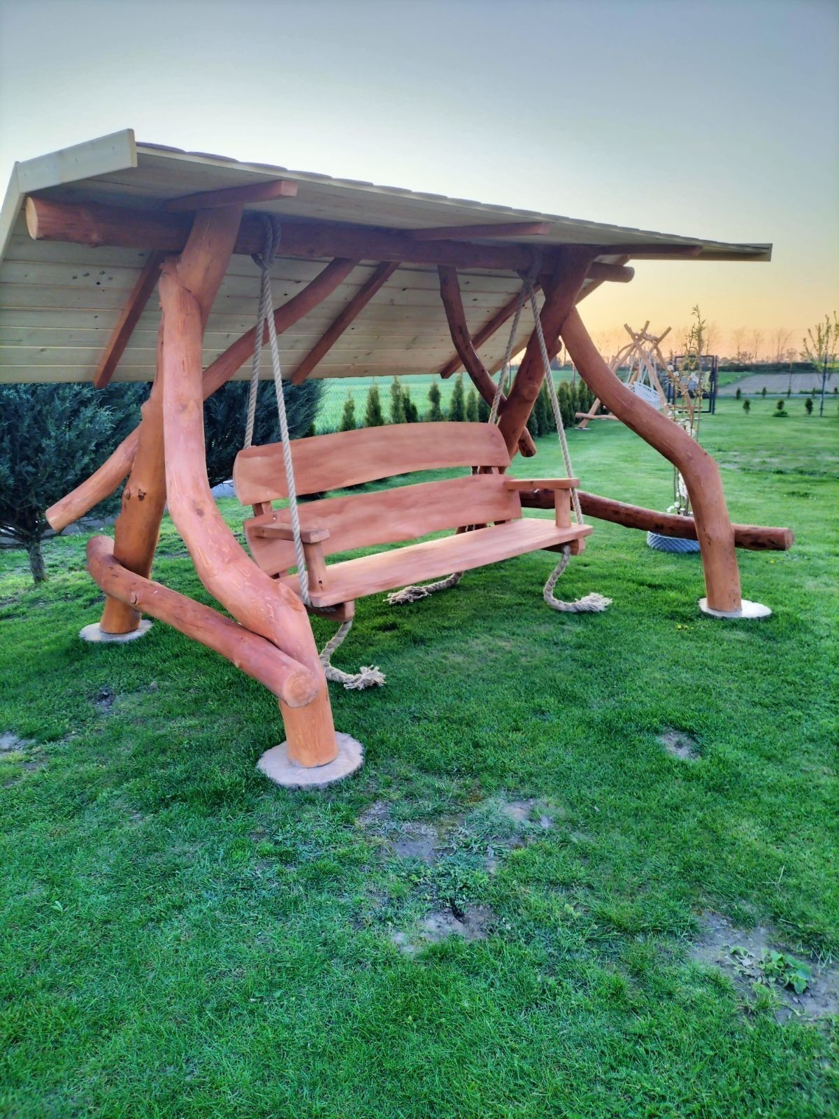 Drewniana huśtawka ogrodowa z zadaszeniem, wykonana z naturalnych pni, zawieszona na linach, osadzona na betonowych podstawach na trawniku, w tle widoczne inne huśtawki i drzewa.