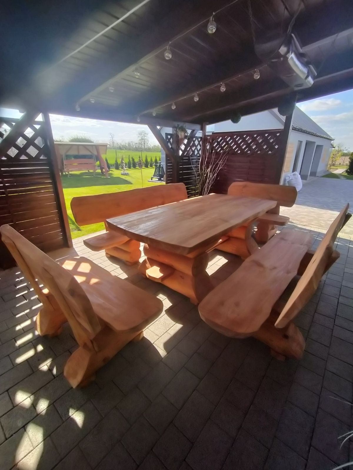 Masywny stół i ławy z surowego drewna pod zadaszeniem z dekoracyjnymi lampkami, w tle widoczny ogród i budynek.