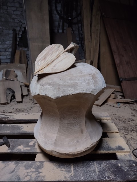 Niedokończona drewniana rzeźba w kształcie owocu z liściem, leżąca na palecie w warsztacie.