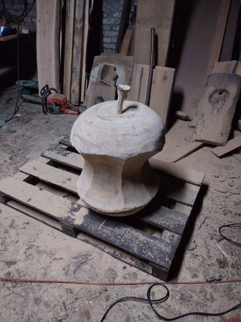 Drewniany, surowy element dekoracyjny przypominający grzyb, umieszczony na palecie w warsztacie, z widoczną piłą łańcuchową w tle.