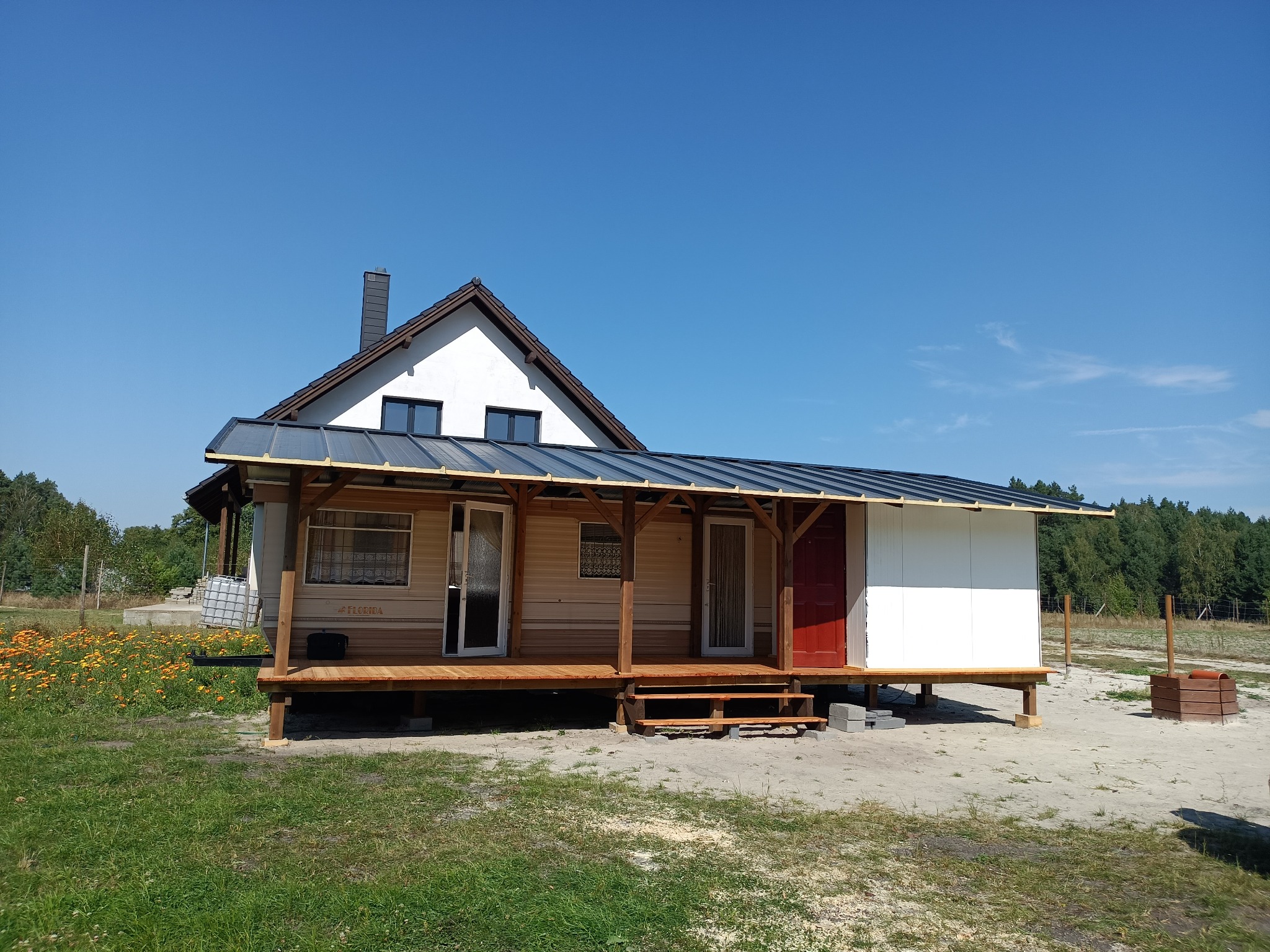 Dom z dobudowanym drewnianym tarasem zadaszonym blachą, widoczne schody i czerwone drzwi wejściowe, na trawniku kwiaty.