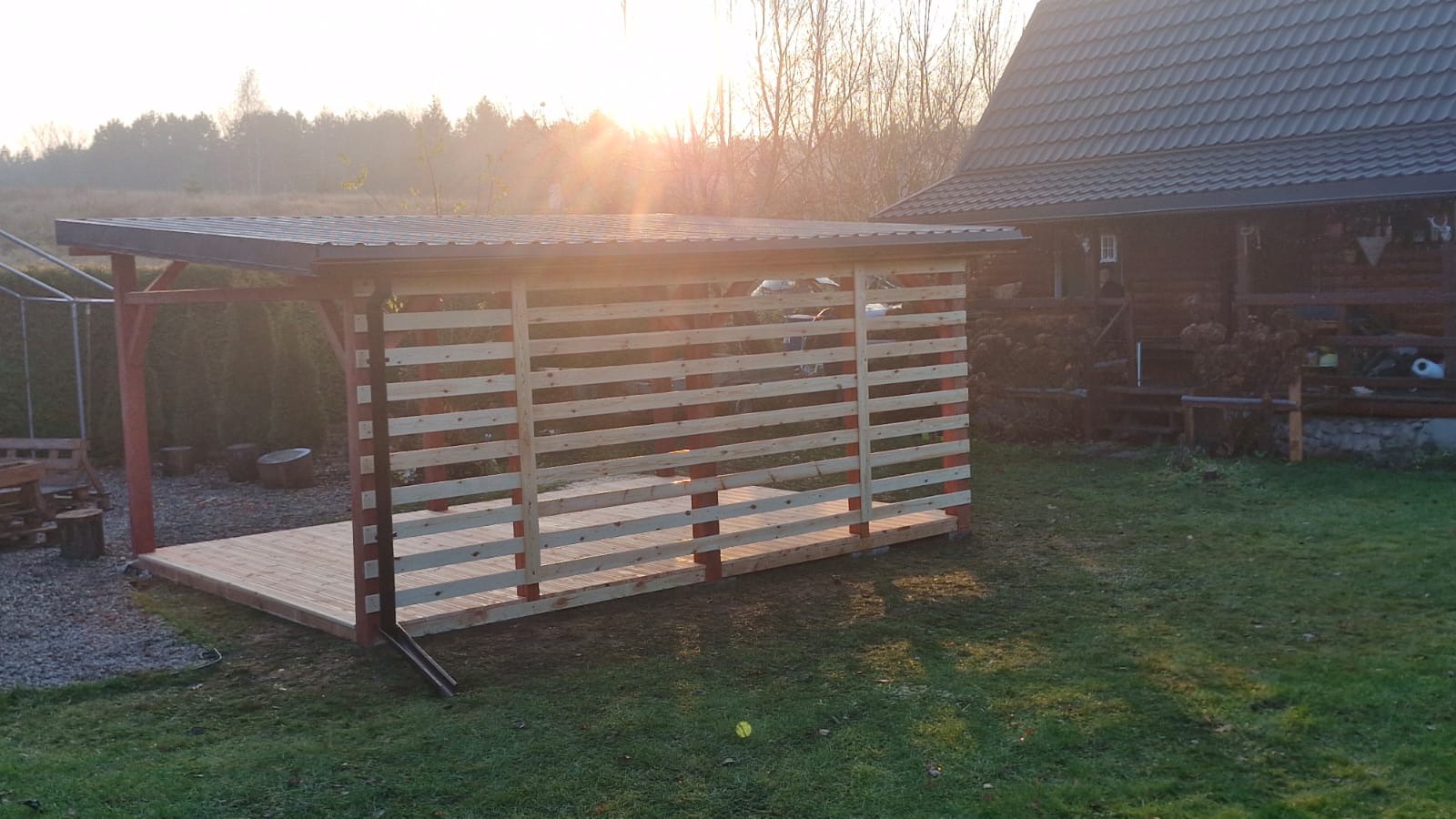 Nowa, częściowo osłonięta konstrukcja z drewnianym podestem i poziomymi listwami, z metalowym dachem, w ogrodzie oświetlonym przez wschodzące słońce.