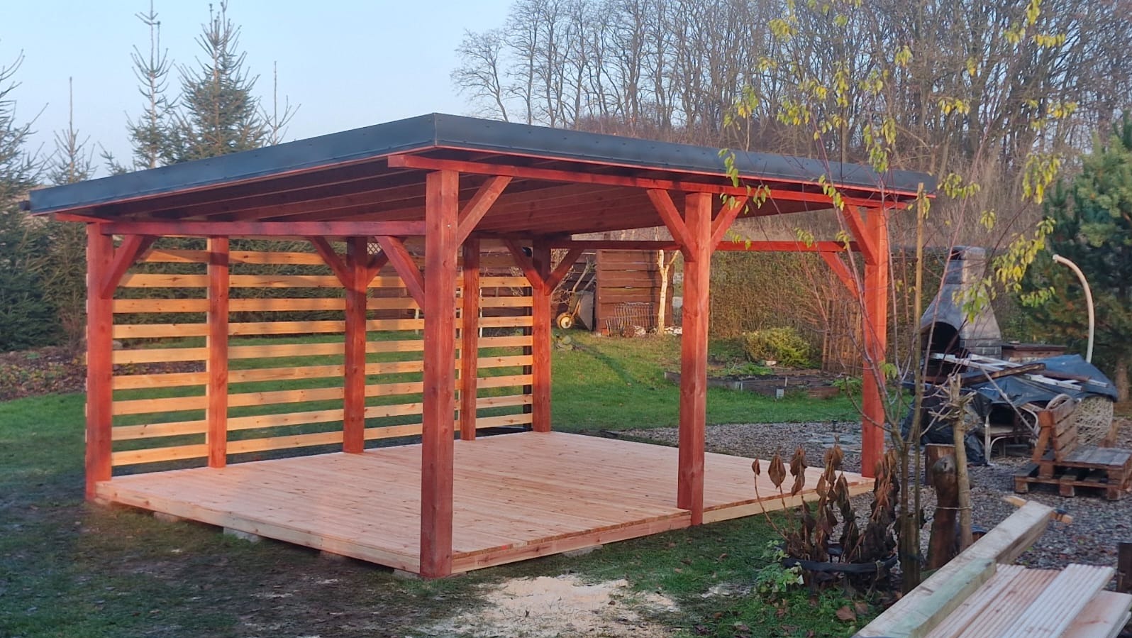 Nowoczesna, otwarta konstrukcja z drewna w kolorze czerwieni, z ażurowymi ścianami i płaskim dachem, na tle ogrodu z grillem.