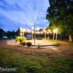 Drewniany podest otoczony piaskiem i dekoracyjnym oświetleniem w ogrodzie o zmierzchu, z widoczną roślinnością i pojazdami w tle.