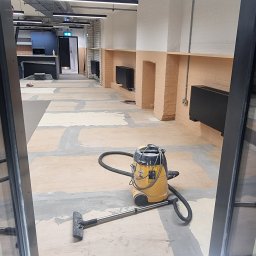 Lukasz Pewinski - Żółty odkurzacz przemysłowy na podłodze z widocznymi śladami po szpachlowaniu, w tle fragment wykończonego pomieszczenia z czarnymi grzejnikami i ceglanymi wnękami.