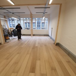 Lukasz Pewinski - Nowo ułożona, jasna podłoga panelowa w pustym pomieszczeniu z oknami i widocznym pracownikiem sprawdzającym coś na telefonie, w tle narzędzia i worki.