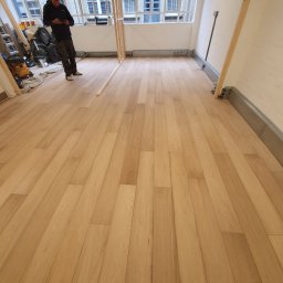 Lukasz Pewinski - Nowo ułożona, jasna podłoga panelowa w przestronnym, jasnym pokoju z dużymi oknami; widoczny pracownik sprawdzający coś na telefonie i przygotowane narzędzia w tle.