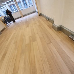 Lukasz Pewinski - Jasne panele podłogowe ułożone w pustym pomieszczeniu, widoczny pracownik w tle i gniazdka elektryczne przy listwach przypodłogowych.