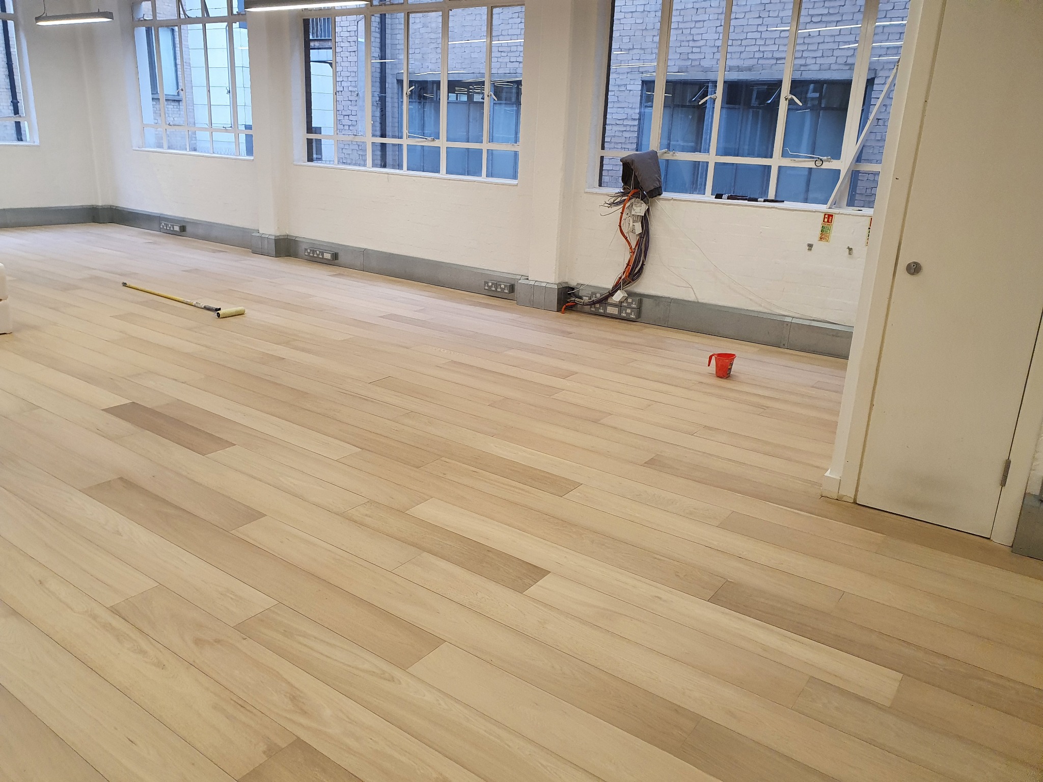 Jasna, drewniana podłoga ułożona w jodełkę w przestronnym, jasnym pomieszczeniu z dużymi oknami; wałek do malowania podłóg leży na podłodze, a czerwony kubek stoi przy ścianie.