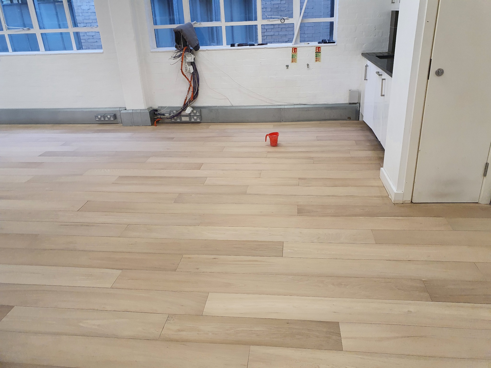 Jasne panele podłogowe w nowym mieszkaniu, widoczne gniazdka elektryczne, okno z niebieskimi zasłonami i instalacja elektryczna wymagająca wykończenia.