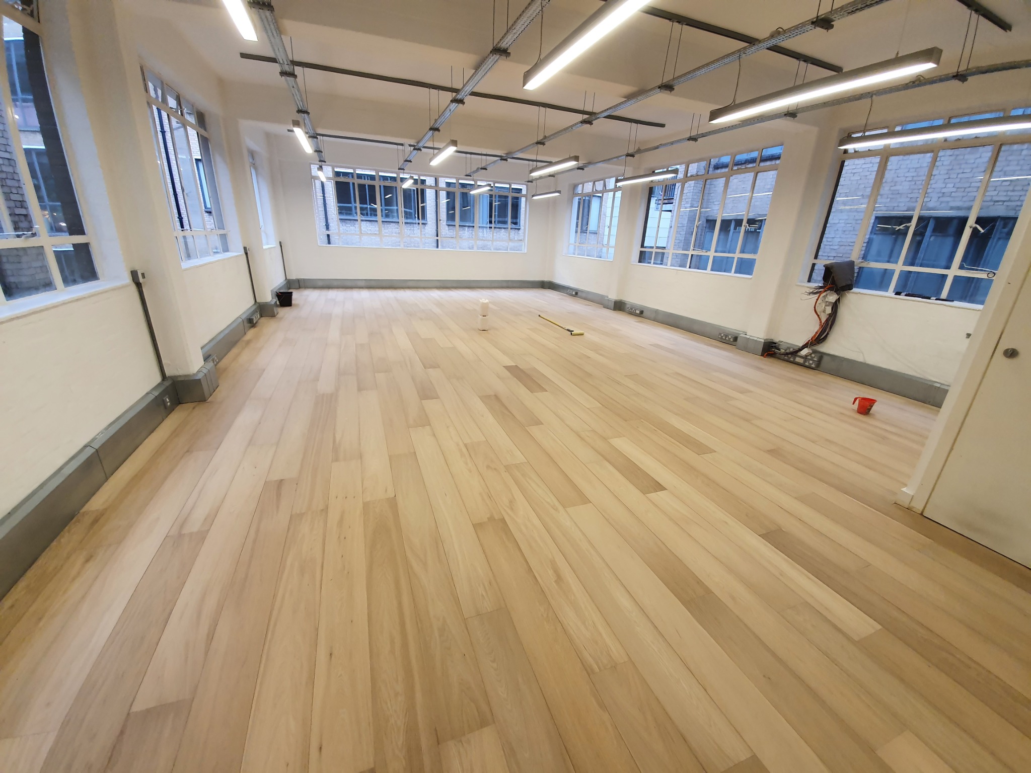 Jasne, drewniane panele podłogowe w przestronnym, jasnym pomieszczeniu biurowym z dużymi oknami i metalowymi lampami sufitowymi.