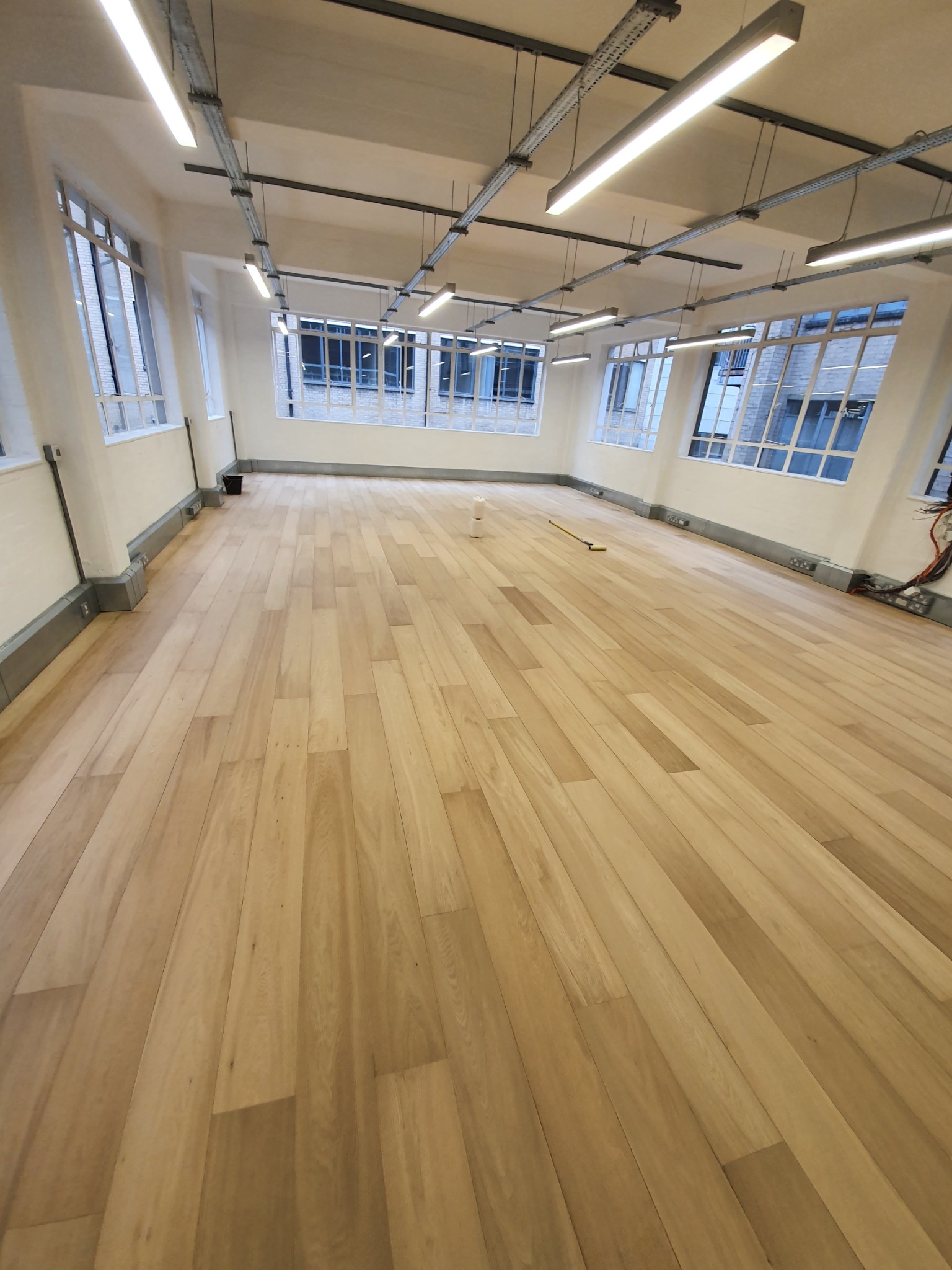 Jasne, drewniane panele podłogowe ułożone w przestronnym, pustym pomieszczeniu z dużymi oknami i przemysłowym oświetleniem sufitowym.