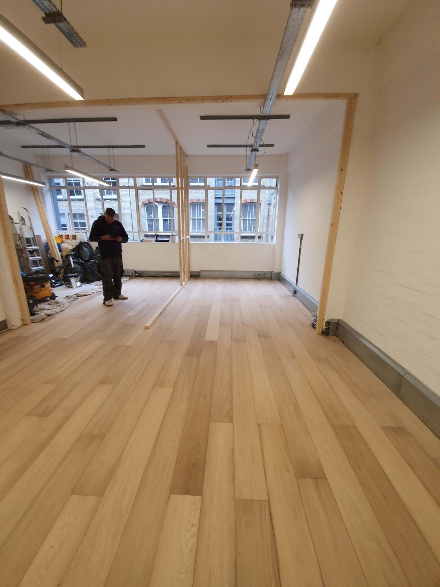 Nowo ułożona, jasna podłoga panelowa w pustym pomieszczeniu z oknami i widocznym pracownikiem sprawdzającym coś na telefonie, w tle narzędzia i worki.
