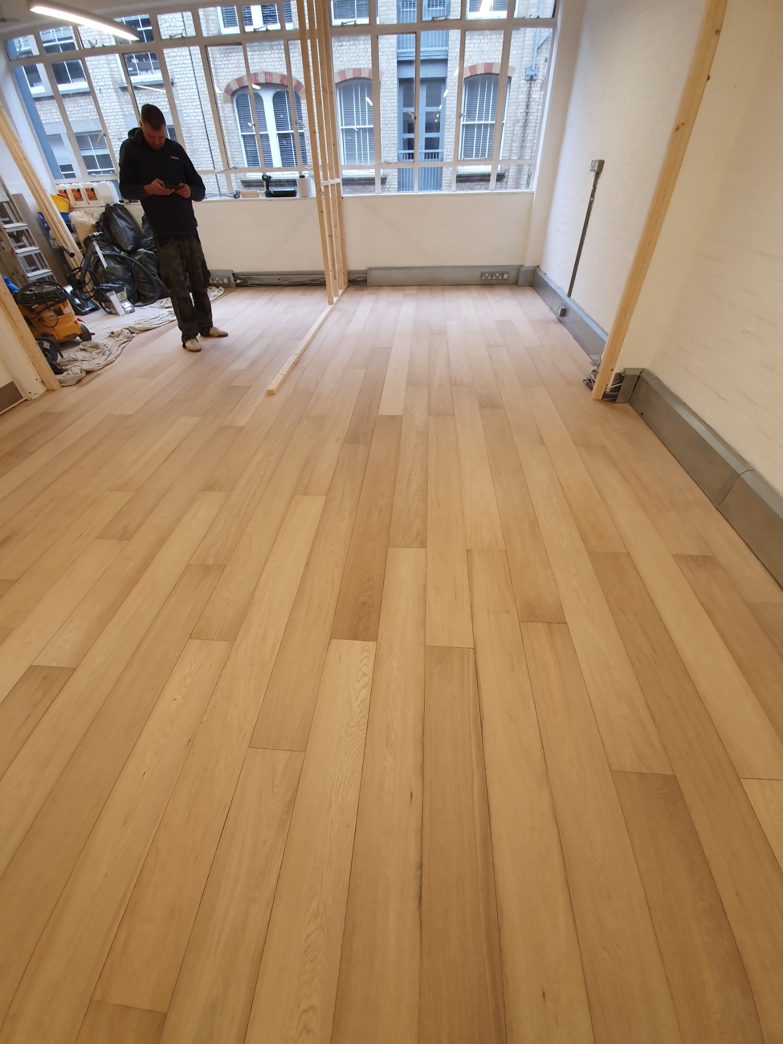 Nowo ułożona, jasna podłoga panelowa w przestronnym, jasnym pokoju z dużymi oknami; widoczny pracownik sprawdzający coś na telefonie i przygotowane narzędzia w tle.