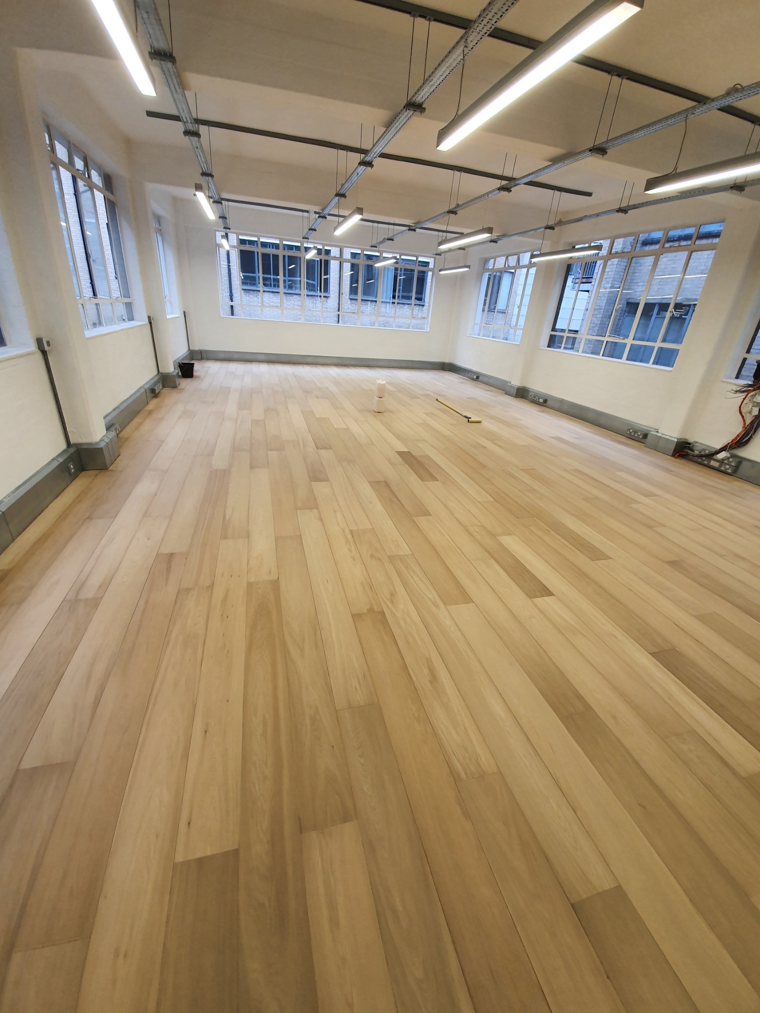 Jasna, drewniana podłoga z paneli w przestronnym, biurowym wnętrzu z rzędami okien i oświetleniem sufitowym, widoczne ślady po wałku malarskim i puszka farby.