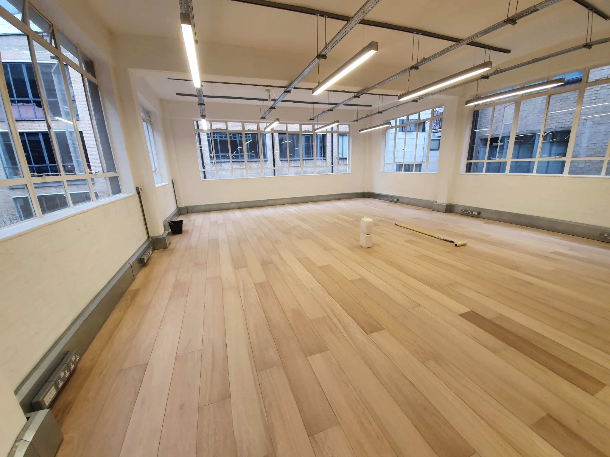 Jasne panele podłogowe w przestronnym, odnowionym pomieszczeniu z dużymi oknami, widoczne gniazdka elektryczne przy podłodze i wałek do malowania.