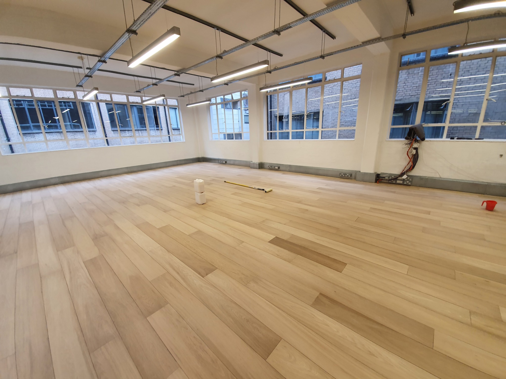 Jasne, nowo położone panele podłogowe w przestronnym pomieszczeniu z dużymi oknami, widoczne narzędzia i materiały przygotowane do dalszych prac wykończeniowych.