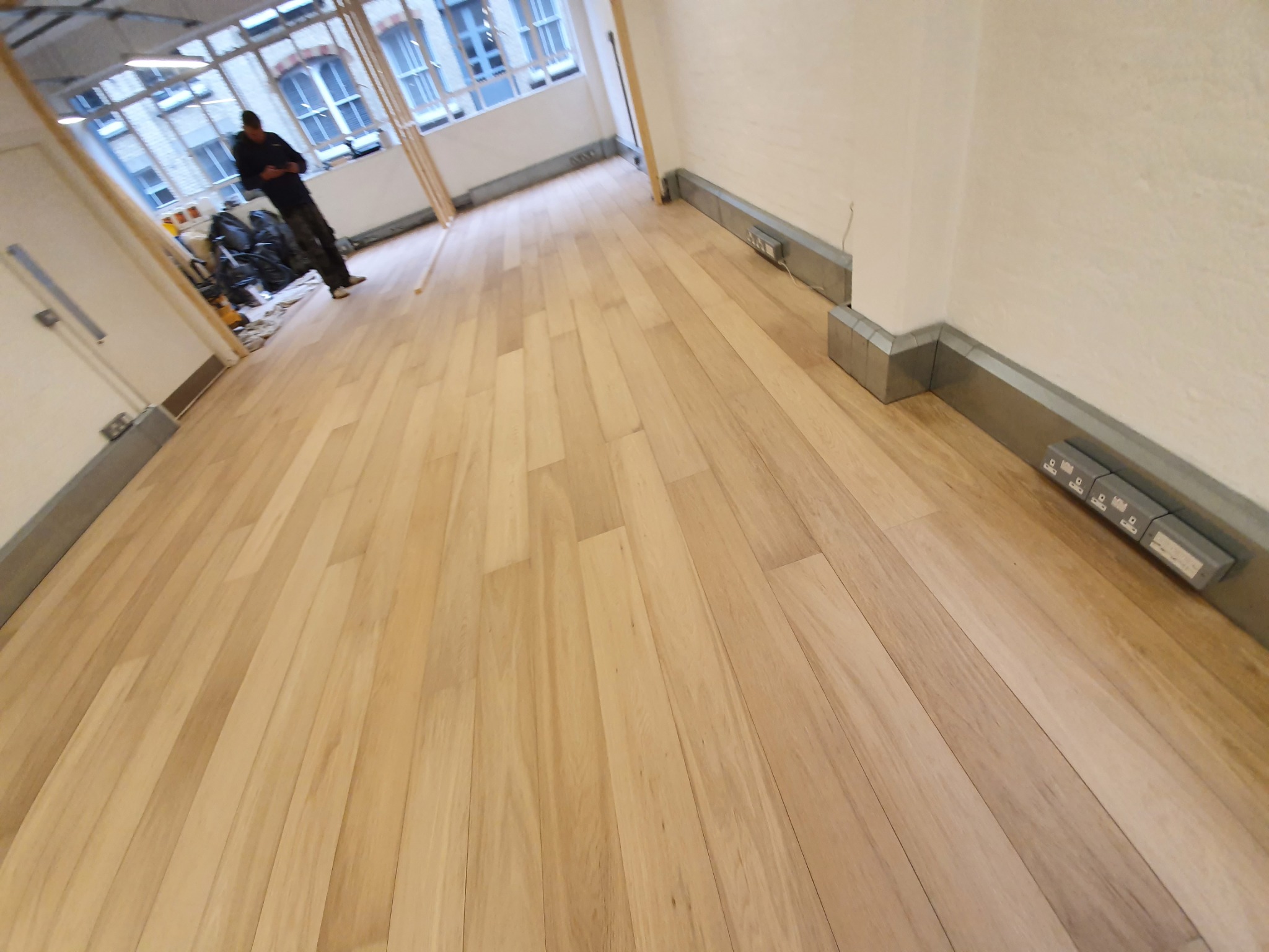 Jasne panele podłogowe ułożone w pustym pomieszczeniu, widoczny pracownik w tle i gniazdka elektryczne przy listwach przypodłogowych.