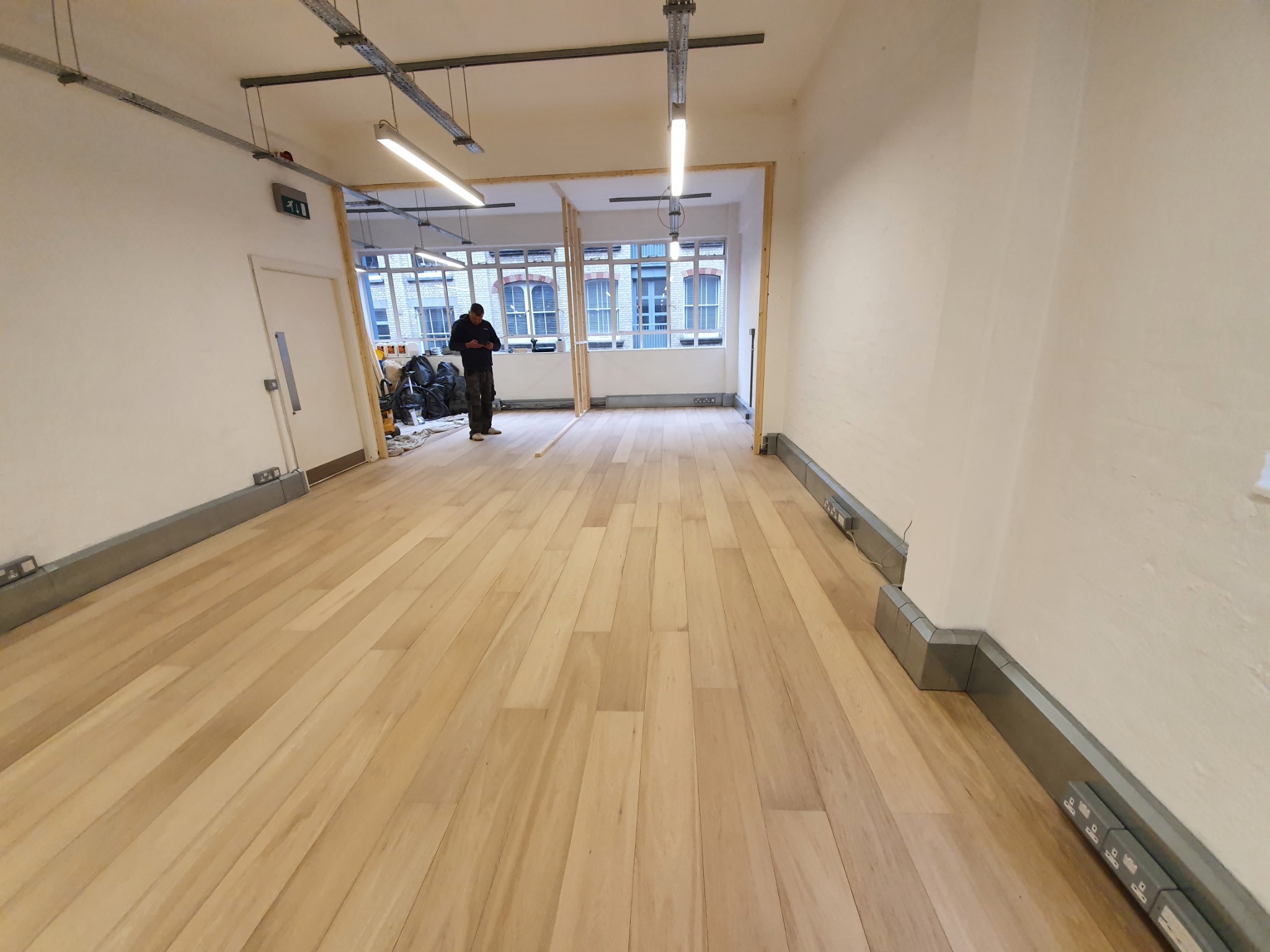 Jasne panele podłogowe ułożone w pustym pomieszczeniu z białymi ścianami, widoczne instalacje elektryczne pod sufitem i pracownik w tle.