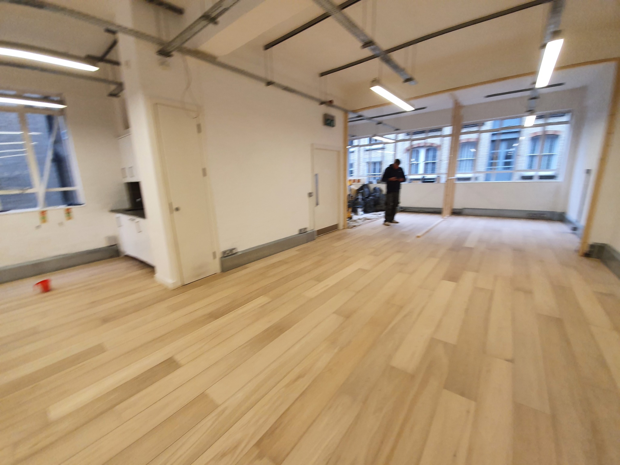 Jasne panele podłogowe w nowo wykończonym, przestronnym pomieszczeniu biurowym z widocznymi instalacjami pod sufitem i osobą w tle.