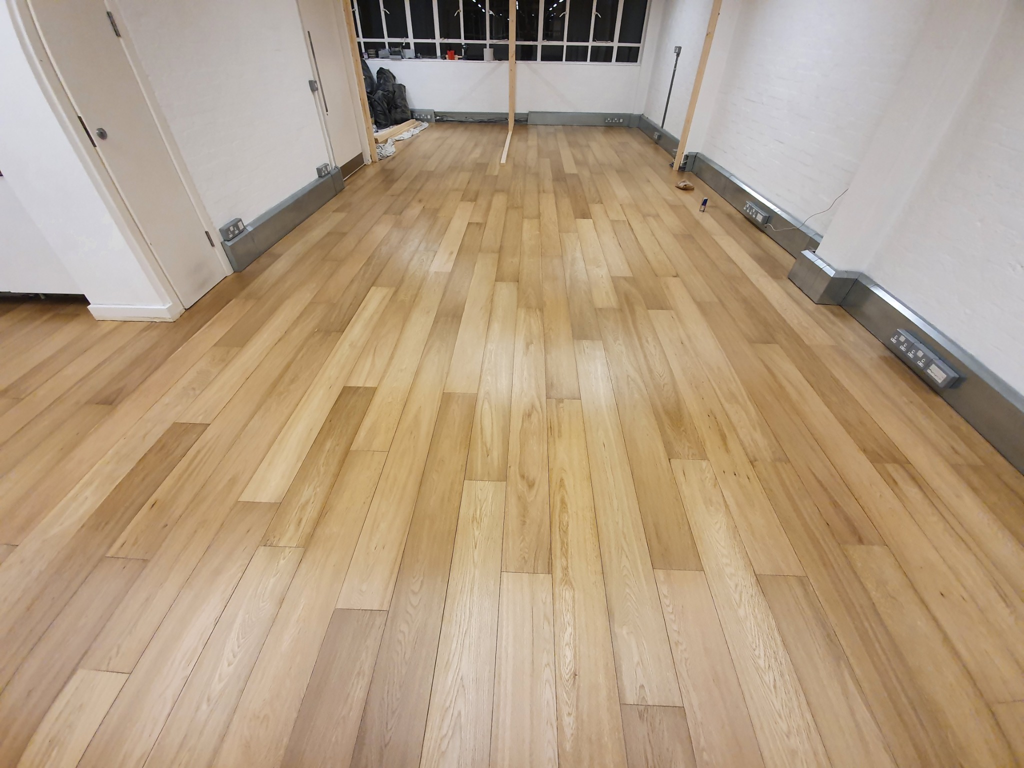 Ukończona podłoga z jasnych paneli drewnianych w pustym pokoju z białymi ścianami i szarymi listwami przypodłogowymi, widoczne gniazdka elektryczne i fragment okna z roletami.