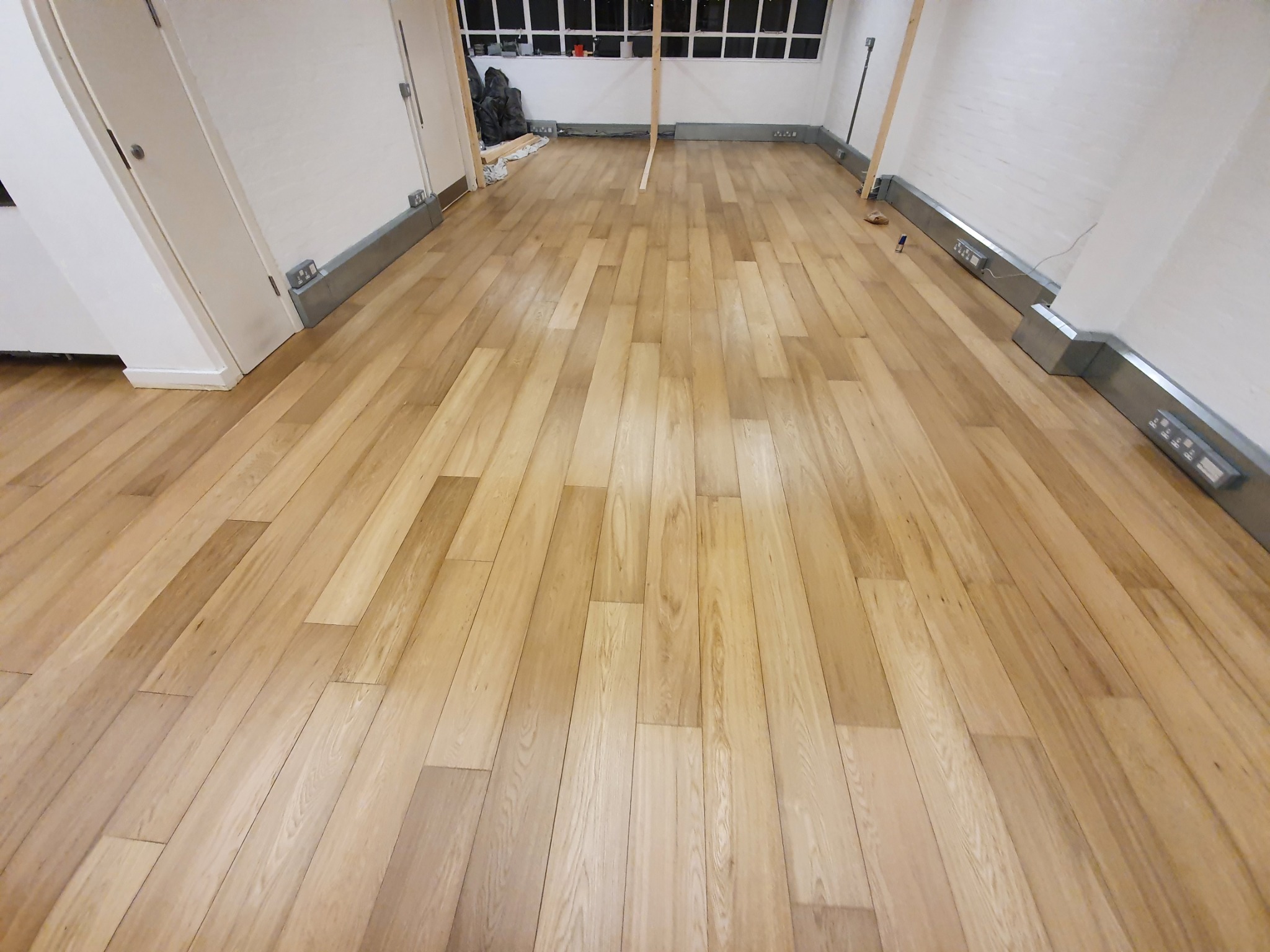 Jasne panele podłogowe ułożone w nowym pomieszczeniu z białymi ścianami i szarą listwą przypodłogową z gniazdkami elektrycznymi.