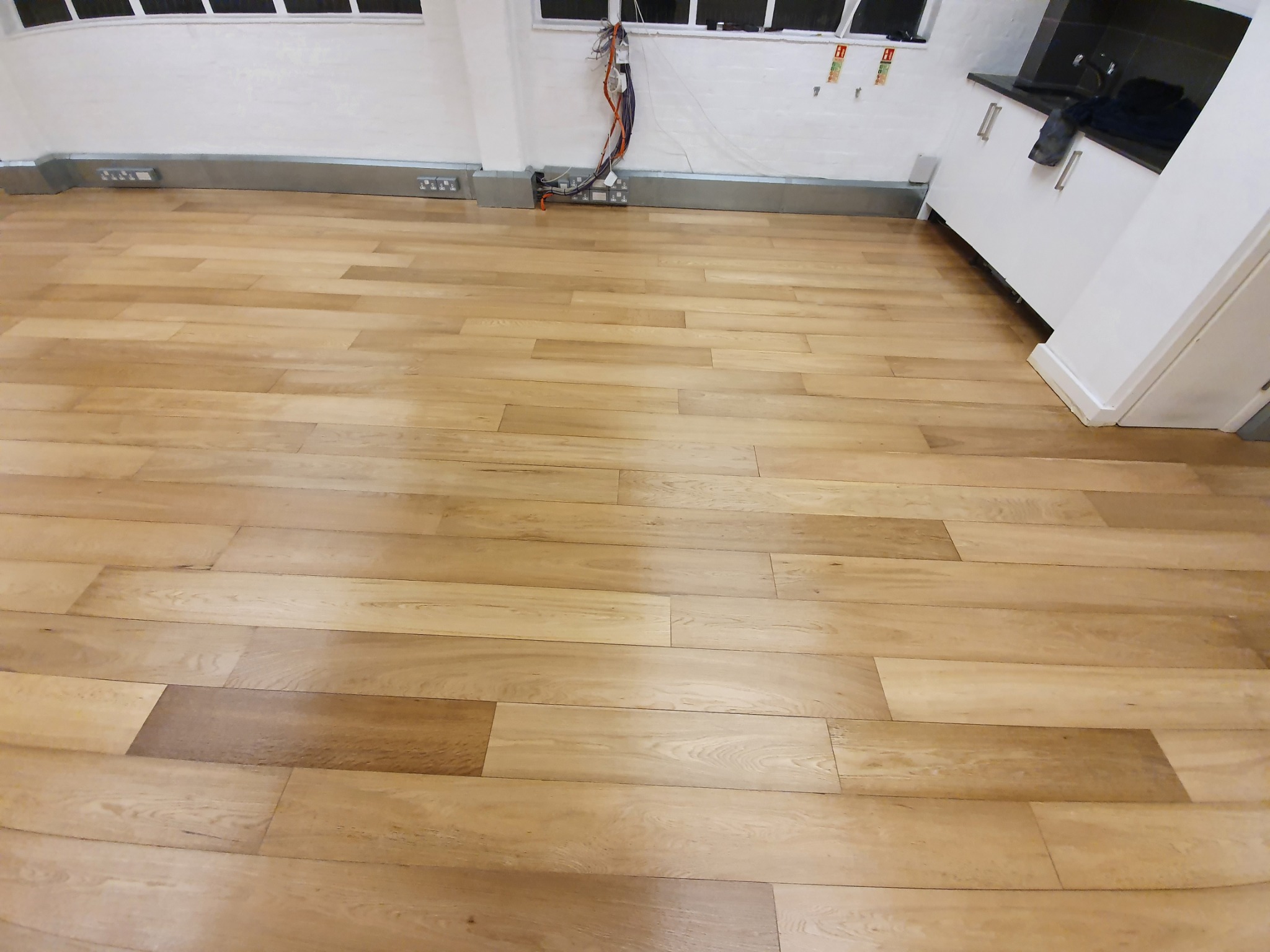 Jasne panele podłogowe ułożone w nowym mieszkaniu, widoczne listwy przypodłogowe i fragment białej zabudowy kuchennej.