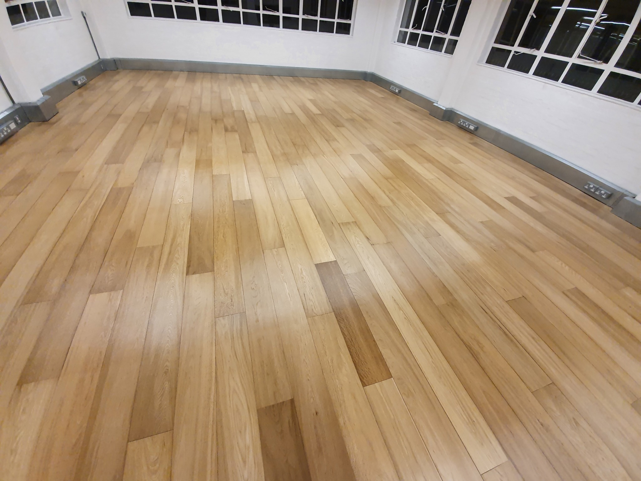 Jasna, świeżo ułożona podłoga z paneli drewnianych, widoczne listwy przypodłogowe i okna w białych ramach.