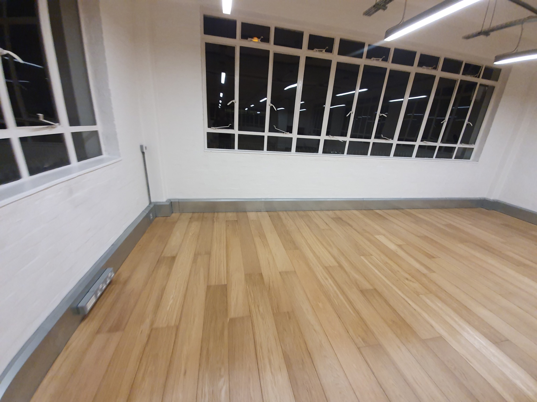 Jasna, drewniana podłoga z listwami przypodłogowymi w odnowionym wnętrzu z dużymi oknami i nowoczesnym oświetleniem.