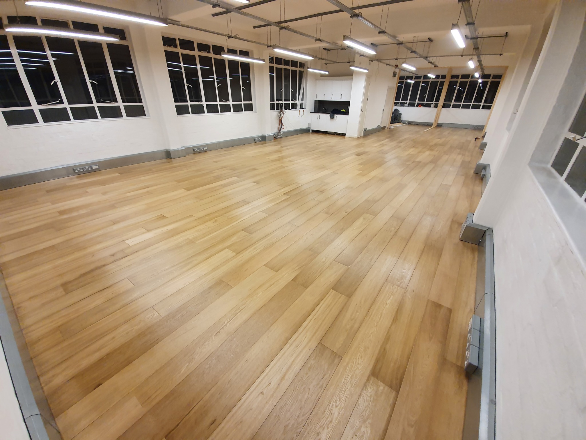 Jasne, drewniane panele podłogowe w przestronnym, odnowionym wnętrzu z dużymi oknami i białymi ścianami, widoczne instalacje sufitowe.