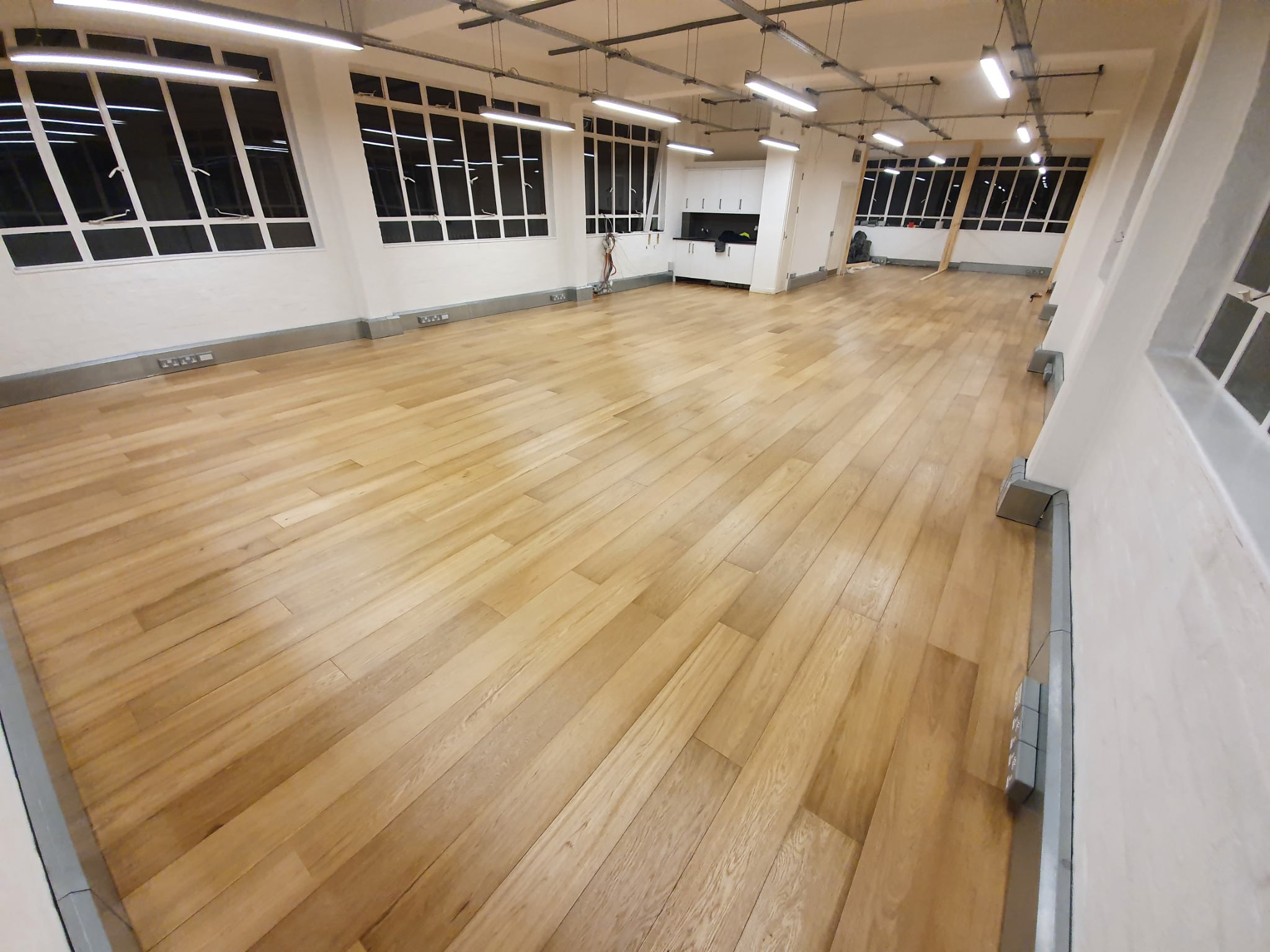 Jasne panele podłogowe ułożone w przestronnym, odnowionym pomieszczeniu z białymi ścianami, dużymi oknami i widocznymi instalacjami sufitowymi.