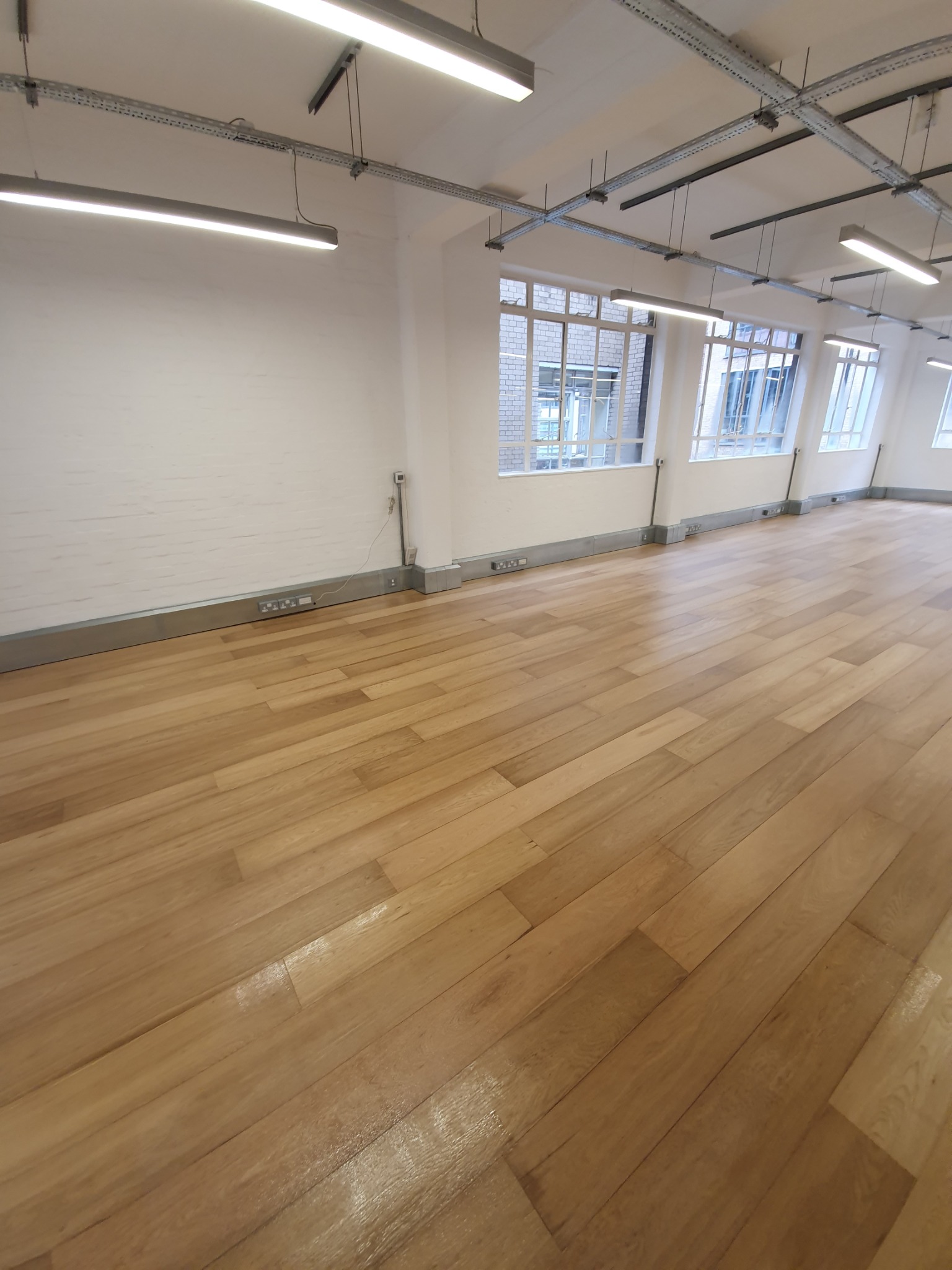 Jasne, błyszczące panele podłogowe w pustym pomieszczeniu z białymi ścianami, oknami i metalowymi elementami pod sufitem.