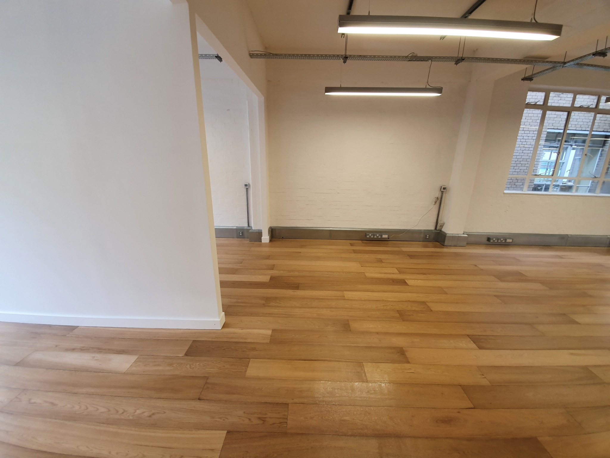 Jasne, drewniane panele podłogowe w pustym, odnowionym pomieszczeniu z białymi ścianami i dużym oknem w tle.