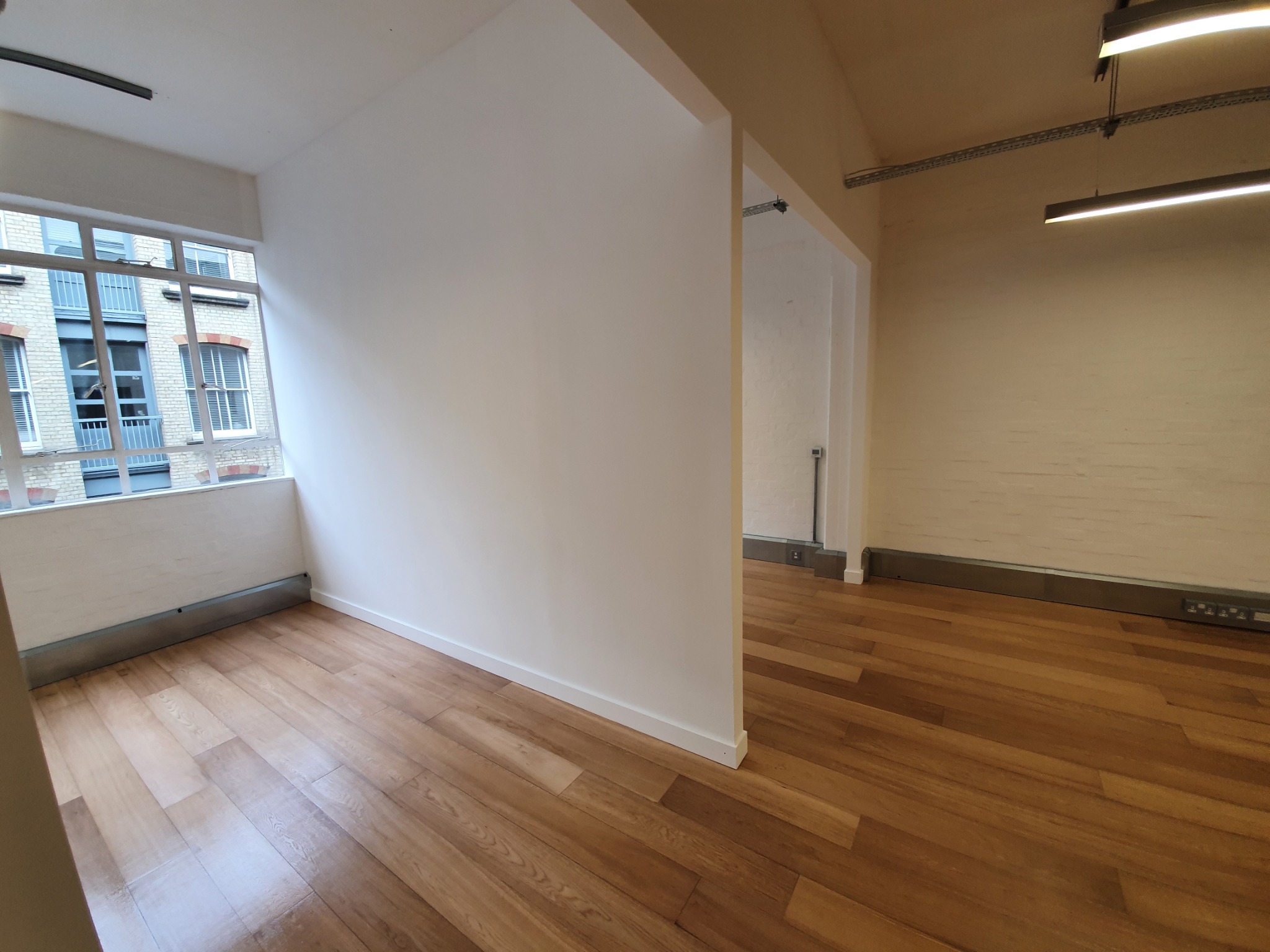 Pomalowane na biało wnętrze z ułożonymi panelami podłogowymi z drewna egzotycznego, widok na okno z zewnętrzną ceglaną zabudową.