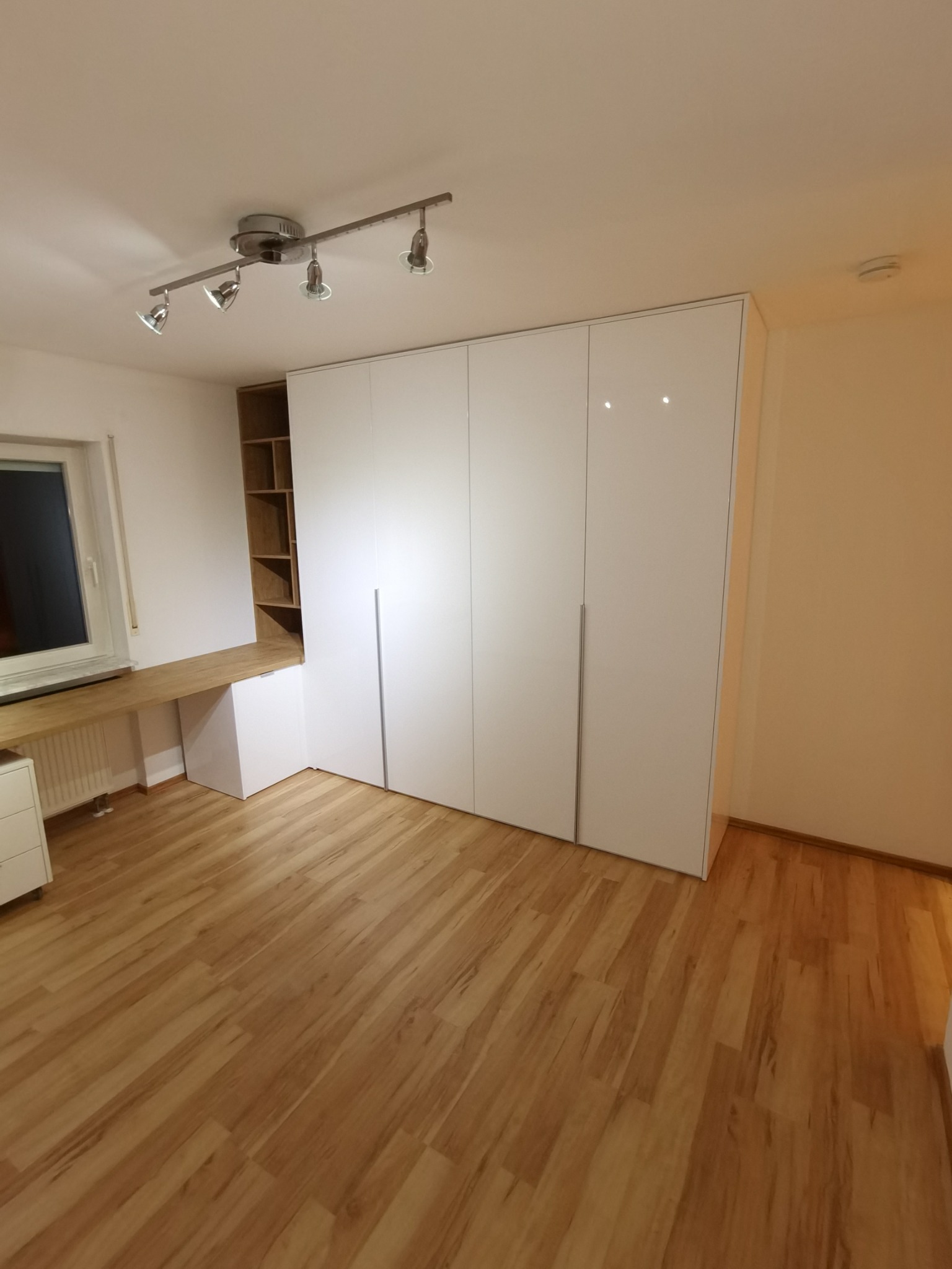 Biała szafa wnękowa z połyskiem, zintegrowana z biurkiem i regałem w pokoju z drewnianą podłogą.