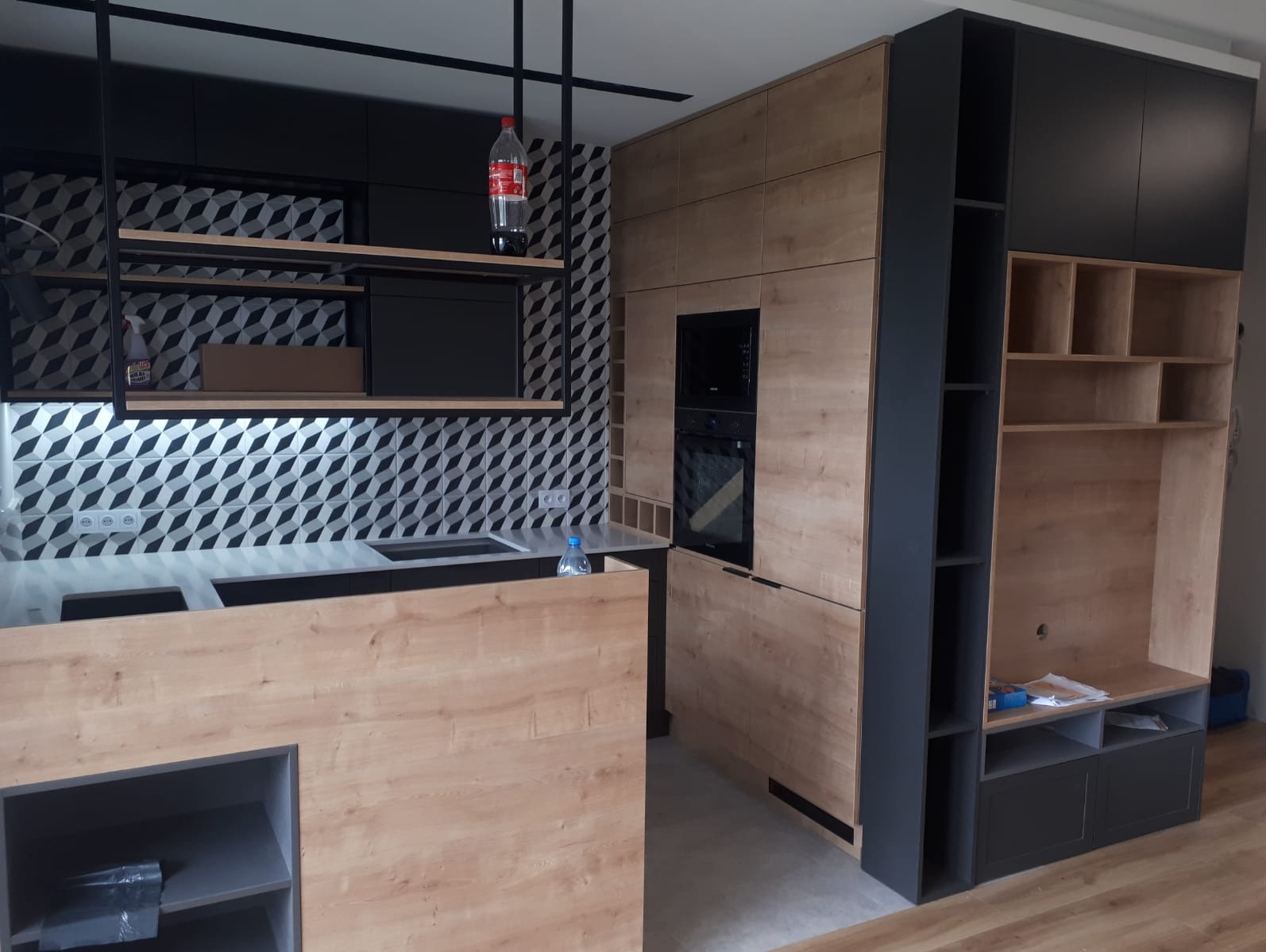 Nowoczesna kuchnia z drewnianymi frontami szafek, czarnymi elementami i geometryczną mozaiką na ścianie, widok na wyspę kuchenną i zabudowany piekarnik.