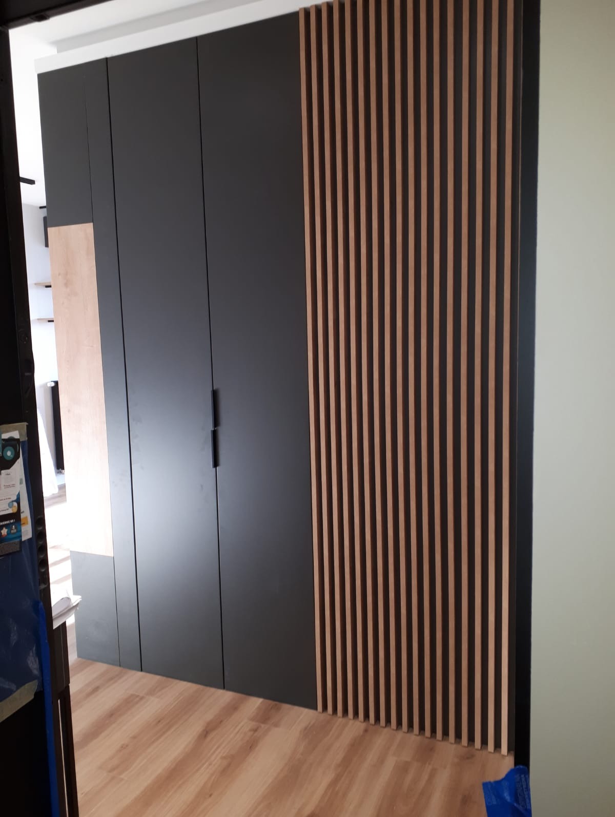 Czarna, nowoczesna szafa z drewnianymi listwami dekoracyjnymi na boku, widoczna z perspektywy podłogi z panelami.