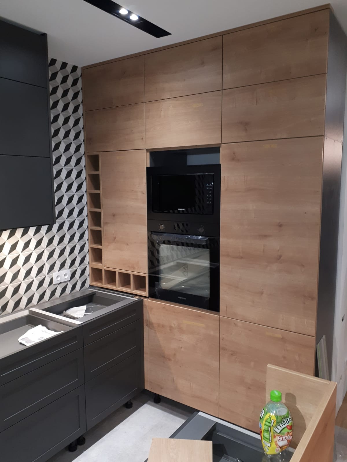 Montaż kuchni na wymiar w Kruszynie: widok na zabudowę z drewnopodobnymi frontami, wbudowany piekarnik i mikrofalówkę oraz szafkę z półkami na wino, na ścianie wzorzyste płytki.