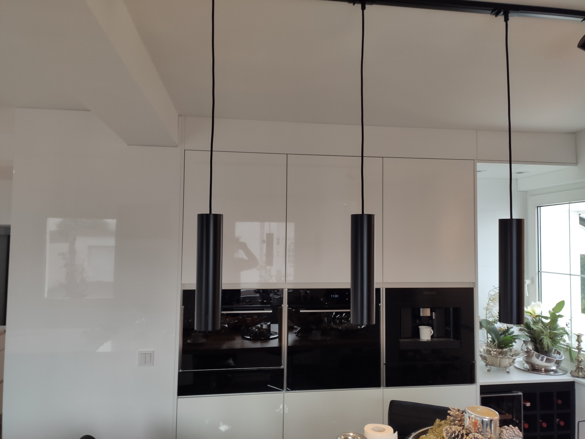 Nowoczesna kuchnia z białymi, lakierowanymi frontami szafek, czarnymi sprzętami AGD w zabudowie i trzema cylindrycznymi lampami wiszącymi na czarnych kablach.