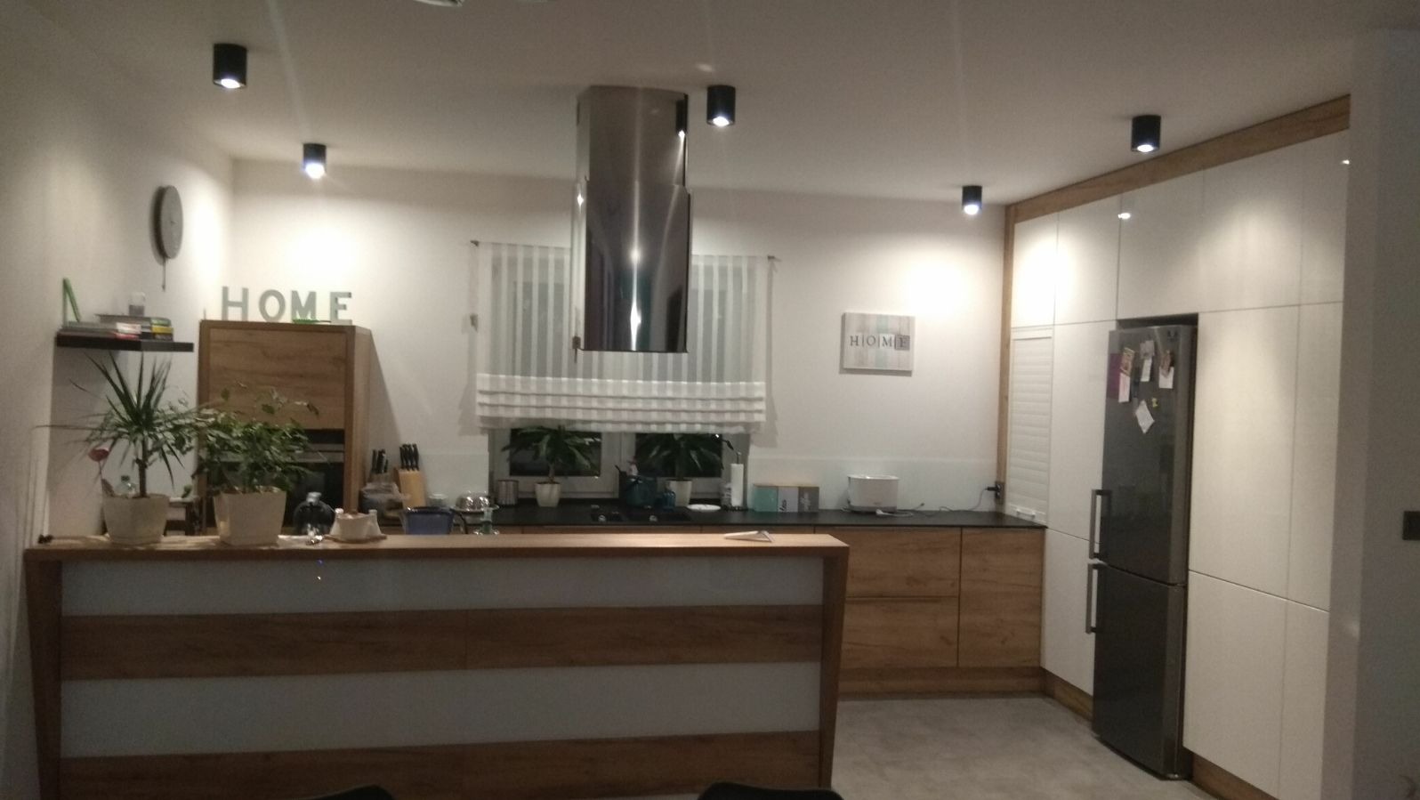 Nowoczesna kuchnia z wyspą, okapem ze stali nierdzewnej, białymi szafkami i drewnianymi akcentami, oświetlona punktowymi lampami sufitowymi.