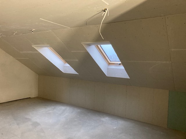 Poddasze z dwoma oknami dachowymi, ściany i sufit wykończone płytami kartonowo-gipsowymi, widoczne przewody elektryczne.
